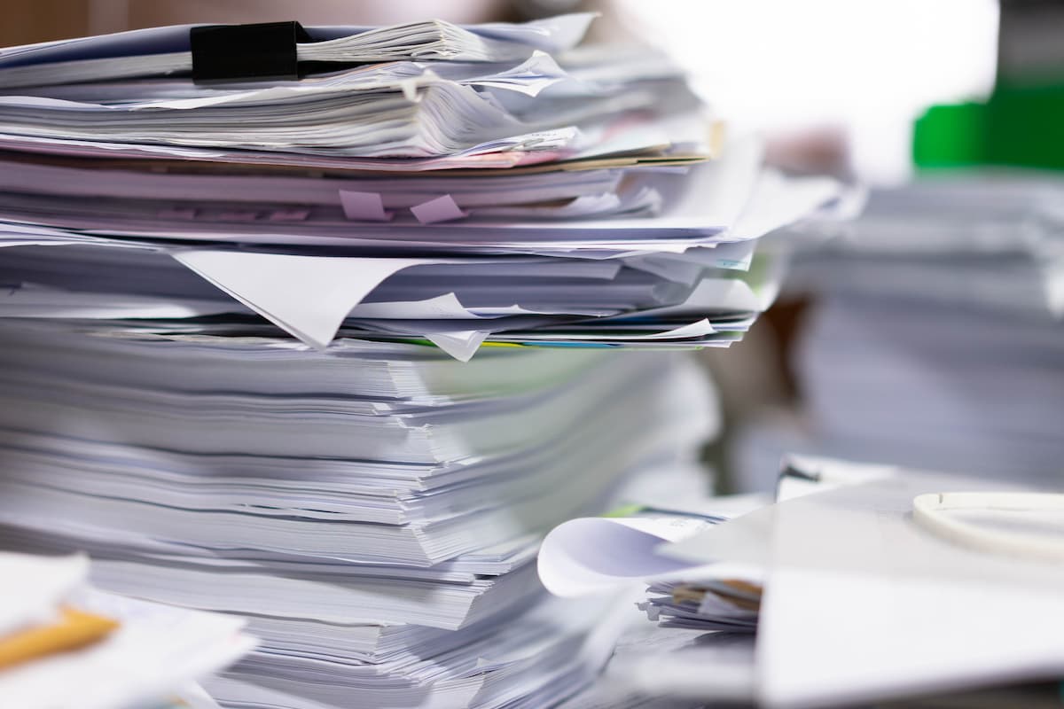業務が特定の担当者に集中し、書類が山積みになった事務所のデスク